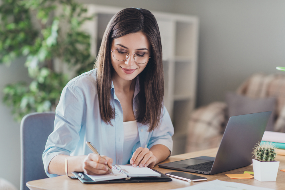 Photo of pretty young girl sit desktop pc hold pen write notepad wear glasses shirt in home office indoors